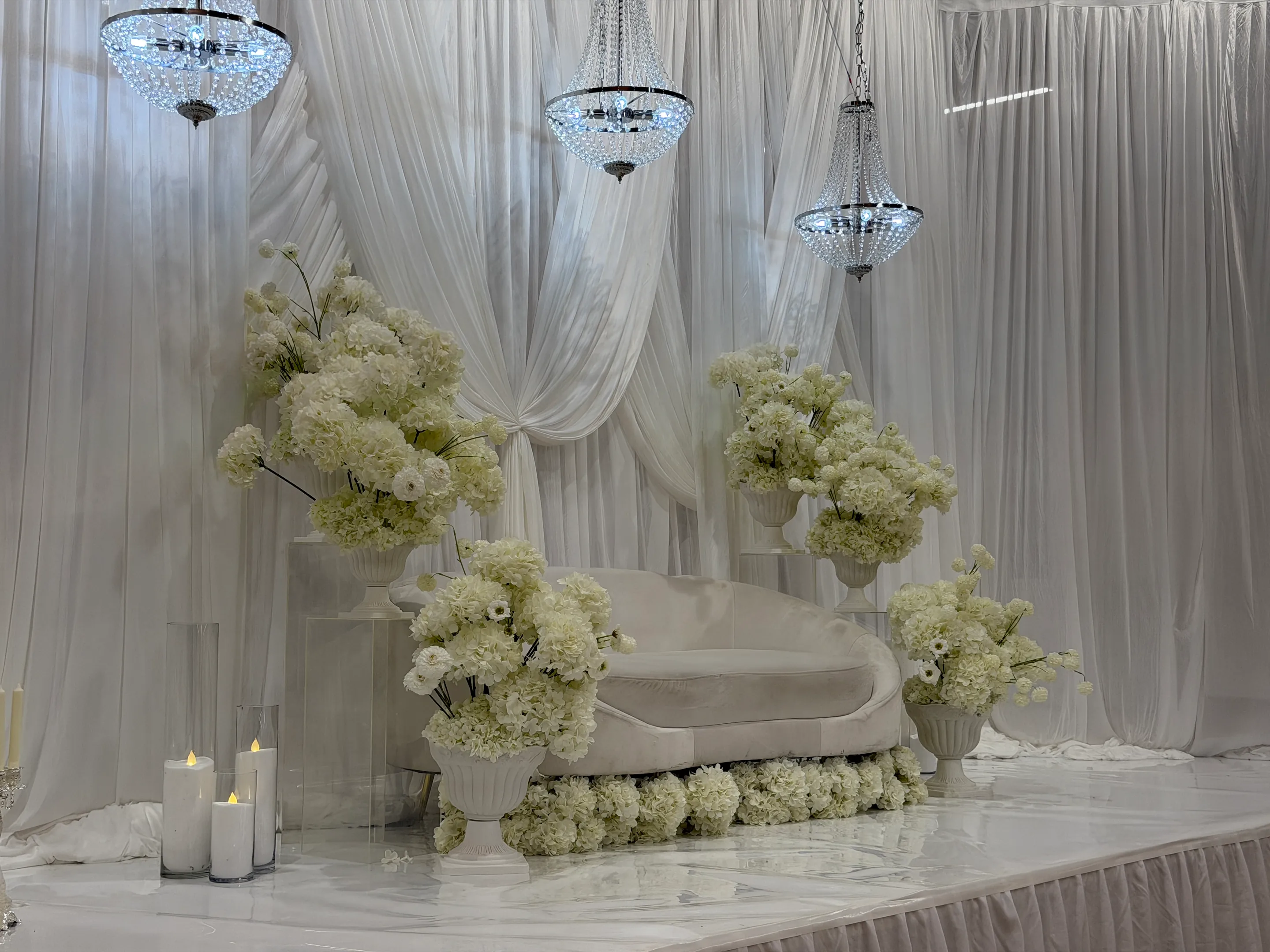 Stage styling by Decoration X: Curved white sofa on a stage edged with white hydrangeas, white florals in urns on acrylic plinths, crystal chandeliers and swagged white drapes, candles in glass.