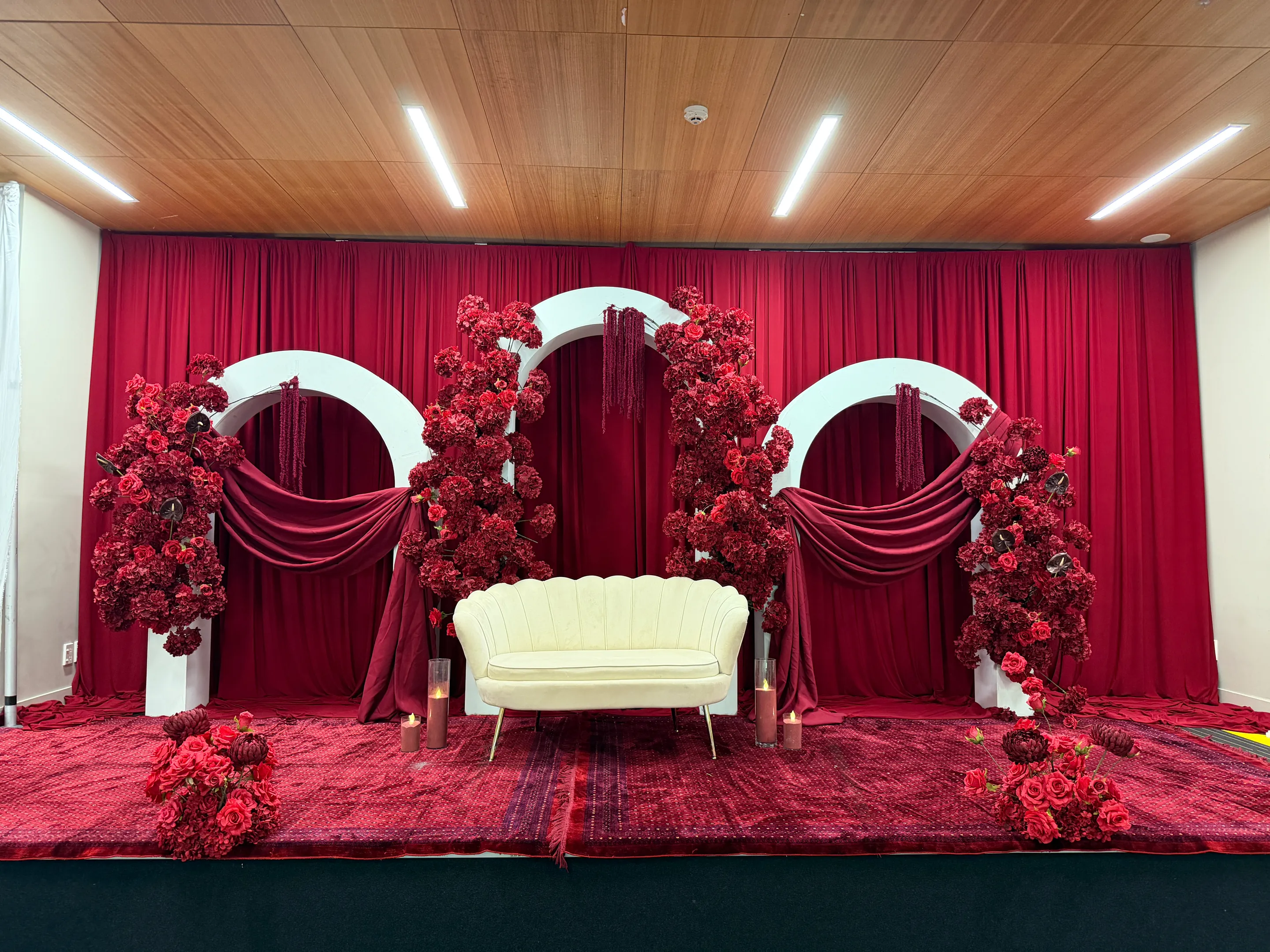 Stage styling by Decoration X: Cream shell-back loveseat on a rug-covered stage, three white arches swathed in burgundy fabric and red rose columns before a deep red curtain, candles in glass.