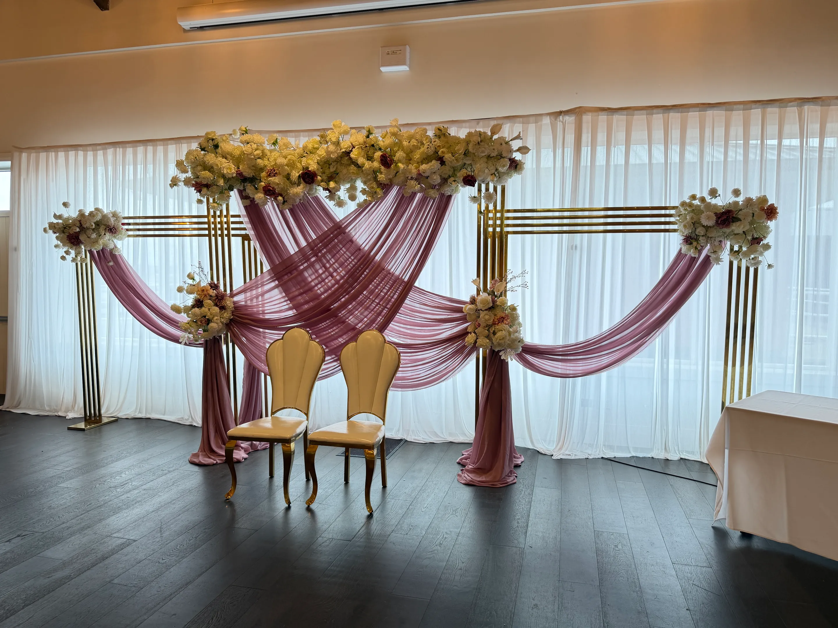 Stage styling by Decoration X: Dusty pink chiffon swagged across gold frame backdrops crowned with cream and burgundy roses, two gold-framed chairs before sheer white curtains.