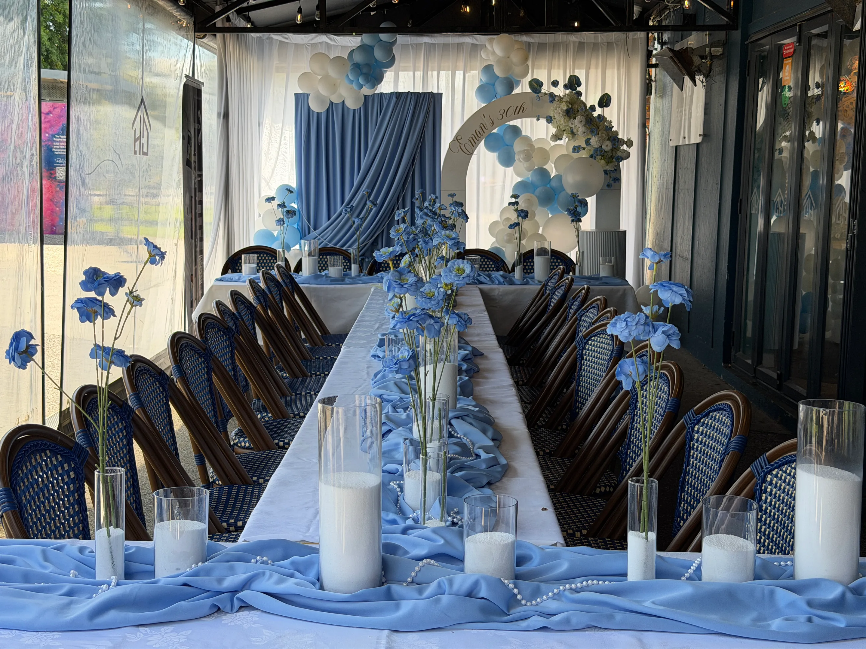 Birthday styling by Decoration X: Long table down a covered terrace dressed in blue chiffon, sand-filled cylinders and blue poppy stems, leading to a blue drape and balloon arch backdrop.