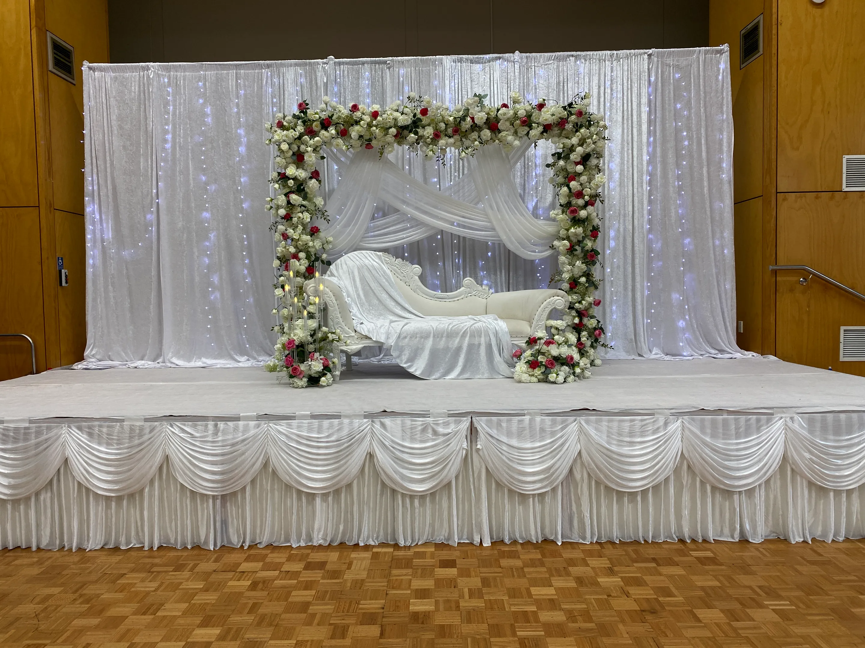 Stage styling by Decoration X: Stage with ruched white skirting and fairy-lit white drapes, a white and red rose square arch over crossed swags and a white carved sofa.